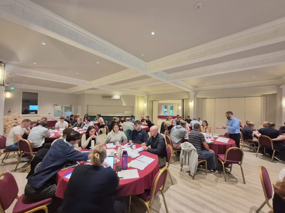 Groups of tables with a man in a blue shirt stood talking to the one table